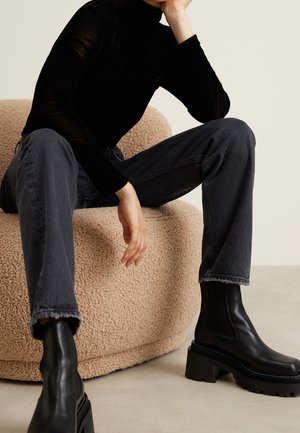 Person sitzt auf einem beige strukturierten Stuhl und trägt einen schwarzen Rollkragenpullover, dunkelgraue Jeans und schwarze klobige Plateau-Stiefel.