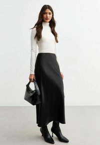 Jupe en satin noir taille haute, associée à un col roulé ajusté blanc. Les accessoires incluent un petit sac à main noir et des bottes noires à talons pointus.