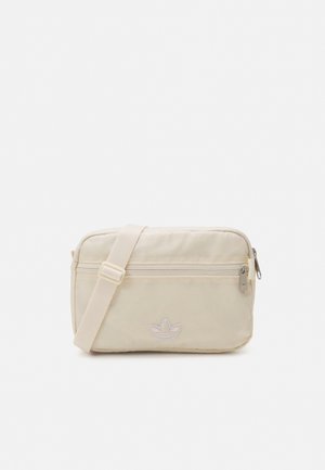 Borsa a tracolla beige con tracolla regolabile, tasca frontale con zip e logo Adidas trefoil discreto su superficie in tessuto liscio.