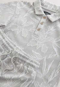 Hellgrauer Pullover mit weißem Blumenmuster und dreiknöpfiger Blende; dazu passende Jogginghose mit elastischem Bund und verstellbarem Kordelzug.