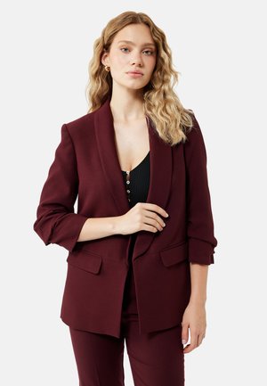 Kvinde med bølget blondt hår iført en burgunderfarvet blazer med op-rullede ærmer og matchende bukser, stående mod en hvid baggrund.
