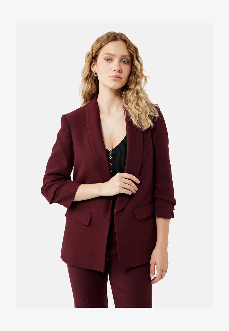 Femme aux cheveux blonds ondulés portant un blazer bordeaux avec les manches retroussées et un pantalon assorti, debout sur un fond blanc.