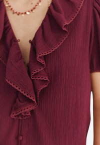 Blusa bordeaux con scollatura arricciata e maniche corte. Il tessuto presenta una finitura testurizzata con un delicato bordo lungo le arricciature e i bottoni.