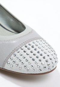 Zapato plano de color gris claro con una parte superior de cuero liso y una puntera texturizada adornada con tachuelas plateadas. Forma redondeada con ribete elástico.