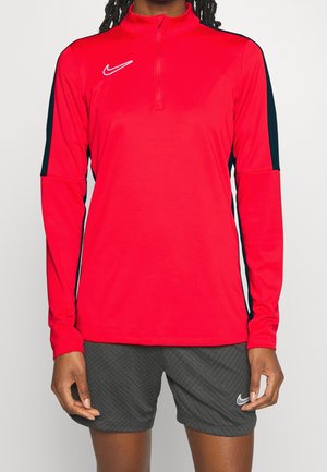 Person iført en rød Nike sportstop med lange ærmer med sorte detaljer og grå Nike-shorts med et subtilt mønster.