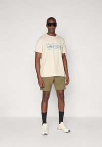 Tricou din bumbac bej cu un design grafic albastru, pantaloni scurți verzi măslinii, șosete negre până la genunchi și adidași deschiși. Fundal simplu.