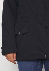 Gros plan d'une personne portant une veste noire avec une poche à boutons-pression et un jean bleu, la main gauche détendue sur le côté.