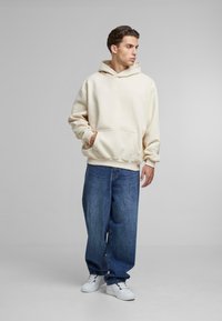 Felpa con cappuccio crema e tasca frontale, abbinata a jeans blu larghi e sneakers bianche. Il tessuto sembra morbido e strutturato.