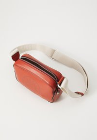 Red leather crossbody bag with a rectangular shape, zip closure, and embossed logo detail. Features a beige fabric strap with lettering.