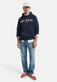 Marineblaues Hoodie mit weißen und burgunderfarbenen Text, mit einer vorderen Tasche, kombiniert mit hellblauen Jeans und roten Sneakers. Weiße Mütze.