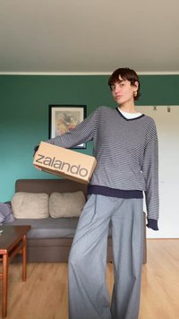 Brauner Karton mit "zalando" in fetter schwarzer Schrift, gehalten von einer Person in einem marineblauen und weißen gestreiften Pullover und grauen, weiten Hosen.