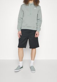 Grå hoodie med dragsnören och framficka, svarta shorts, samt gröna och vita rutiga slip-on skor med vita strumpor.