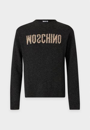 Mężczyzna w czarnym swetrze z odwróconym tekstem "MOSCHINO", niebieskich dżinsach i czarnych butach stoi z rękami w kieszeniach na jednolitym tle.