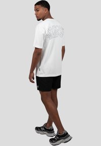 Bílá bavlněná trička s nápisem "WRONG FRIENDS" tištěným tučně na zádech, nošené s černými šortkami a šedými teniskami. Krátké rukávy a volný střih.