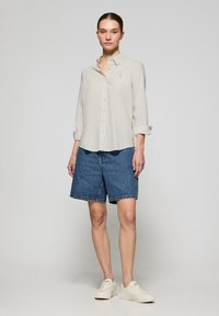 Camicia button-up beige chiaro con colletto, abbinata a pantaloni corti di jeans blu scuro. Il modello indossa sneaker di colore chiaro. Design minimalista.