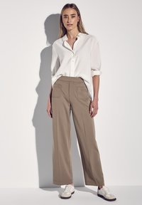 Beiga vida byxor med fickdetaljer, kombinerat med en vit skjorta med knappar. Modellen bär vita och beige sneakers. Slät tygstruktur.