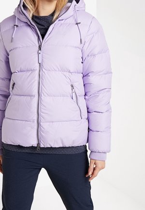 Mujer que lleva una chaqueta acolchada ligera color púrpura claro con capucha, bolsillos con cremallera y puños acanalados, combinada con pantalones oscuros y una blusa negra.