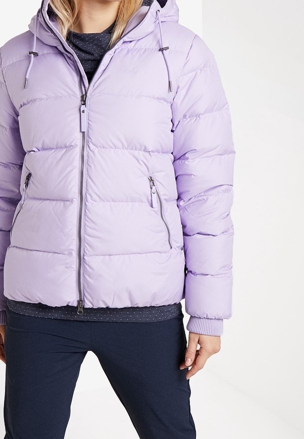 Femme portant une doudoune matelassée légère violet clair avec capuche, poches zippées et poignets côtelés, associée à un pantalon foncé et un haut noir.