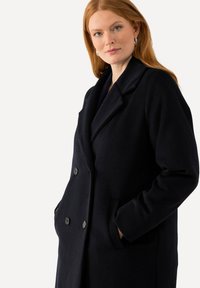 Námořnický vlněný dvouřadý kabát s klopeným límcem, velkými knoflíky a bočními kapsami. Materiál je hladký a texturovaný.