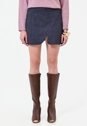 Donna che indossa una minigonna asimmetrica blu scuro, un maglione rosa, braccialetti dorati spessi e stivali di pelle marroni al ginocchio con punte dorate.