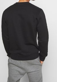 Felpa nera con colletto rotondo, maniche lunghe e polsini a coste. Presenta una texture liscia e un design minimalista, abbinata a pantaloni della tuta grigi.