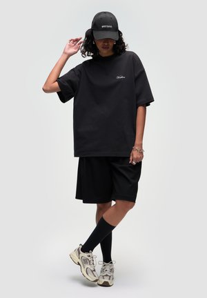 T-shirt oversize nera con logo ricamato, pantaloni corti neri larghi, calze nere fino al ginocchio e scarpe da corsa beige. Outfit casual e sportivo.