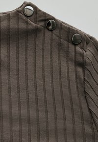 Brauner gerippter Stoff mit vertikalen Streifen, der drei metallische Druckknöpfe an der Schulter zum Verschließen besitzt. Glatte Textur, taillierter Schnitt.