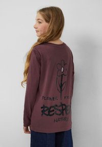 Majica dugih rukava u burgundijskoj boji sadrži veliki crni cvjetni grafikon i tekst "CVIJET IZ POŠTOVANJA PREMA PRIRODI" na poleđini. Mekani pamučni materijal.