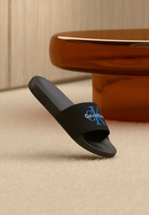 Schwarze Calvin Klein Slide-Sandale mit blauem "CK"-Logo auf dem Riemen, aufrecht stehend auf beigefarbenem Teppich in der Nähe eines runden bernsteinfarbenen Tisches.