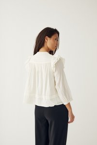 Femme aux longs cheveux foncés portant un chemisier blanc ample avec des épaules volantées et un pantalon noir, de profil sur un fond uni.