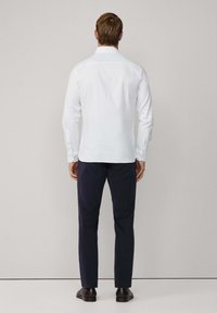Chemise blanche à manches longues avec un col pointu, poignets boutonnés, texture lisse, assortie à un pantalon sombre et des chaussures noires, vue de dos.