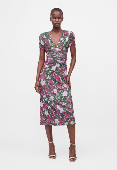 Femme portant une robe fleurie longueur genou avec des fleurs roses et blanches sur fond vert, debout les mains le long du corps.