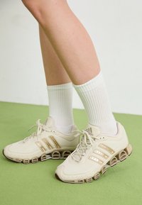 Scarpe sportive beige con tomaia testurizzata, dettagli dorati e suola segmentata evidente. Abbinate a calzini bianchi a coste.