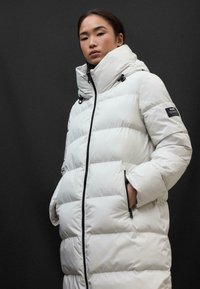 Piumino bianco con colletto alto, trapuntatura orizzontale, zip nera e tasche laterali. Presenta cordini sul cappuccio e un'etichetta con il marchio.