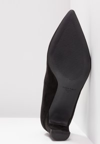 Chaussure noire à bout pointu avec semelle texturée, taille 37, posée en angle contre un mur d'angle blanc sur une surface plane.