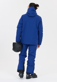 Blå skidjacka med huva, i kombination med matchande blå byxor. Håller en svart hjälm och har svarta stövlar accentuerade och slitstarkt material.