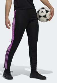 Persona che indossa pantaloni da tuta neri con strisce viola che tiene accanto a sé un pallone da calcio Adidas bianco e nero.