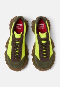 Helle grüne und braune Wanderschuhe mit strukturierten Sohlen und Schnürsenkeln, die ein rotes und weißes Logoetikett innen aufweisen.