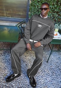 Siva kapuca in trenirke z belimi obrobki, v kombinaciji s sijočimi črnimi čevlji. Model sedi za zeleno zunanjo mizo na kameniti podlagi.