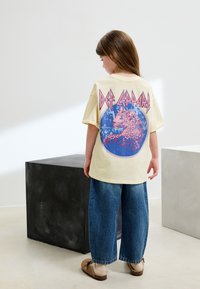 Gelbes übergroßes T-Shirt mit einem grafischen Design eines Leoparden in einem blauen Kreis, darüber pinkfarbener Schriftzug "Def Leppard".