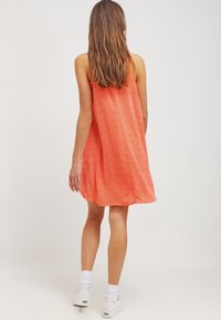 Ausgestelltes A-Linien-Kleid in leuchtendem Orange mit Zickzack-Muster, aus leichtem Stoff gefertigt. Das Modell trägt weiße Sneaker und Knöchelsocken.