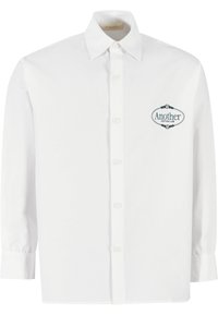 Hvit button-up skjorte laget av bomull, med en spiss krage og et brodert logo på venstre bryst. Langermede med knappedeksel.