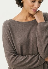 Bruine gebreide trui met een wijde halslijn en een relaxte fit, met lange mouwen en een zacht textuur. Gouden hoop oorbellen als accent.