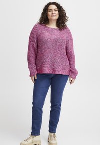 Stickad tröja i rosa med flerfärgade inslag, vida ärmar och en lös passform. Matchas med blå jeans och beigefärgade snörskor.