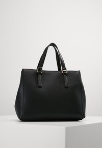 Borsa in pelle nera a granuli con manici superiori doppi, forma strutturata e dettagli hardware in tono oro, appoggiata su una superficie grigia chiara.
