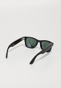 Occhiali da sole in plastica nera con lenti verde scuro. Design con montatura quadrata e superficie liscia. Dettagli in metallo vicino alle cerniere e logo sul lato.