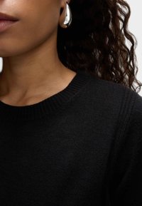 Maglione nero a maglia con colletto a girocollo a coste, abbinato a un orecchino argentato lucido e scultoreo. Lo sfondo è neutro e discreto.