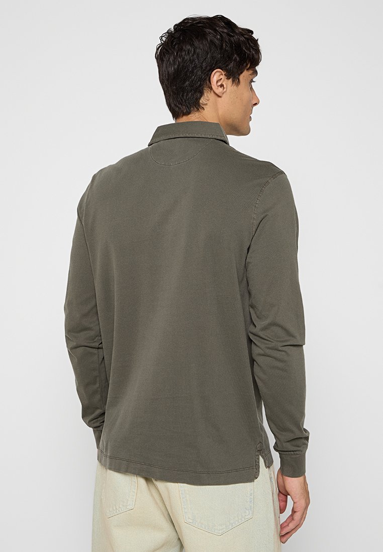 Ein Mann, der von hinten zu sehen ist, trägt ein langärmliges, dunkelolivgrünes Hemd und hellbeige Hosen vor einem schlichten weißen Hintergrund.