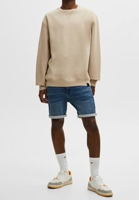 Sweat-shirt beige à encolure ronde, associé à un short en denim bleu avec ourlets roulés. Chaussures blanches à accents orange.