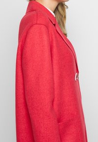 Abrigo de lana rojo con una textura suave, cuello de muesca y cierre de un solo botón. Presenta bolsillos laterales y un ajuste entallado.